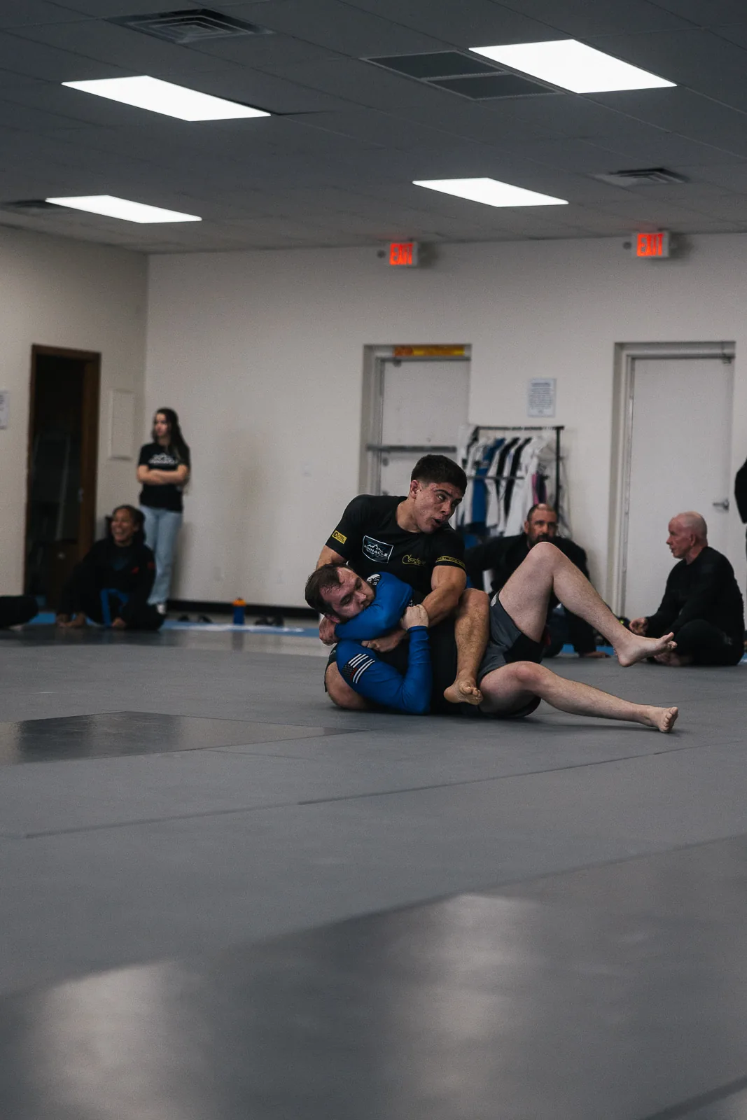 Two adults rolling during a no-gi class while teammates watch from the edge of the mat